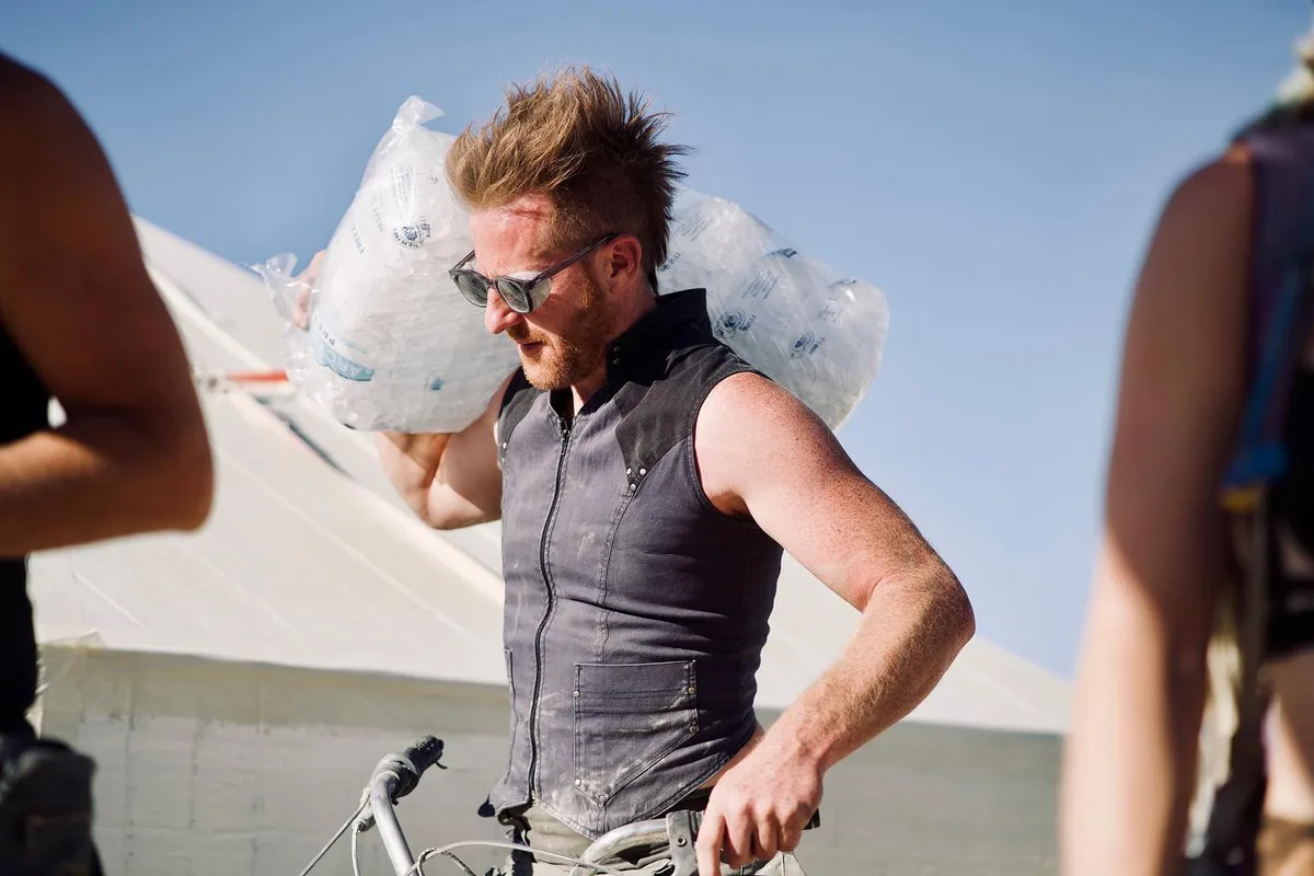 A person carrying clear bags of ice over one shoulder beside a bicycle