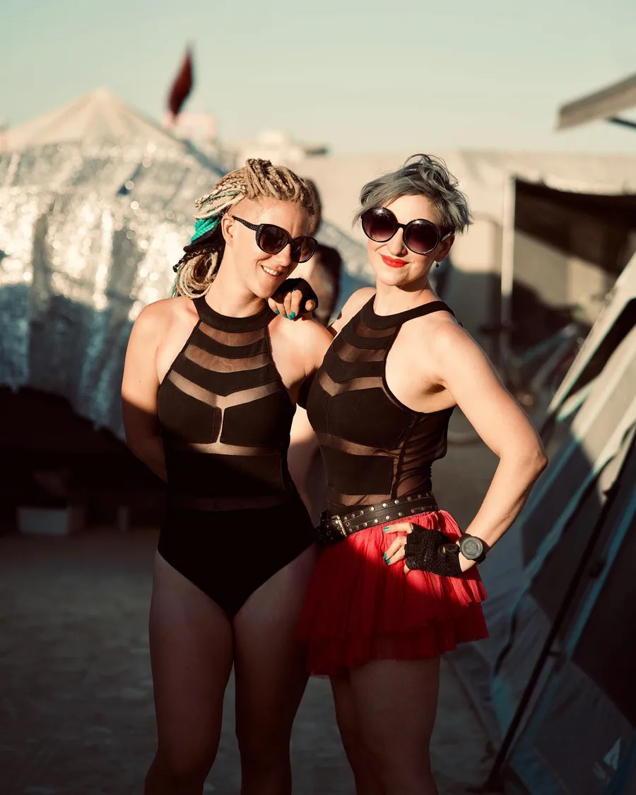 Two people in matching black outfits and sunglasses pose arm in arm, one wearing a red tutu