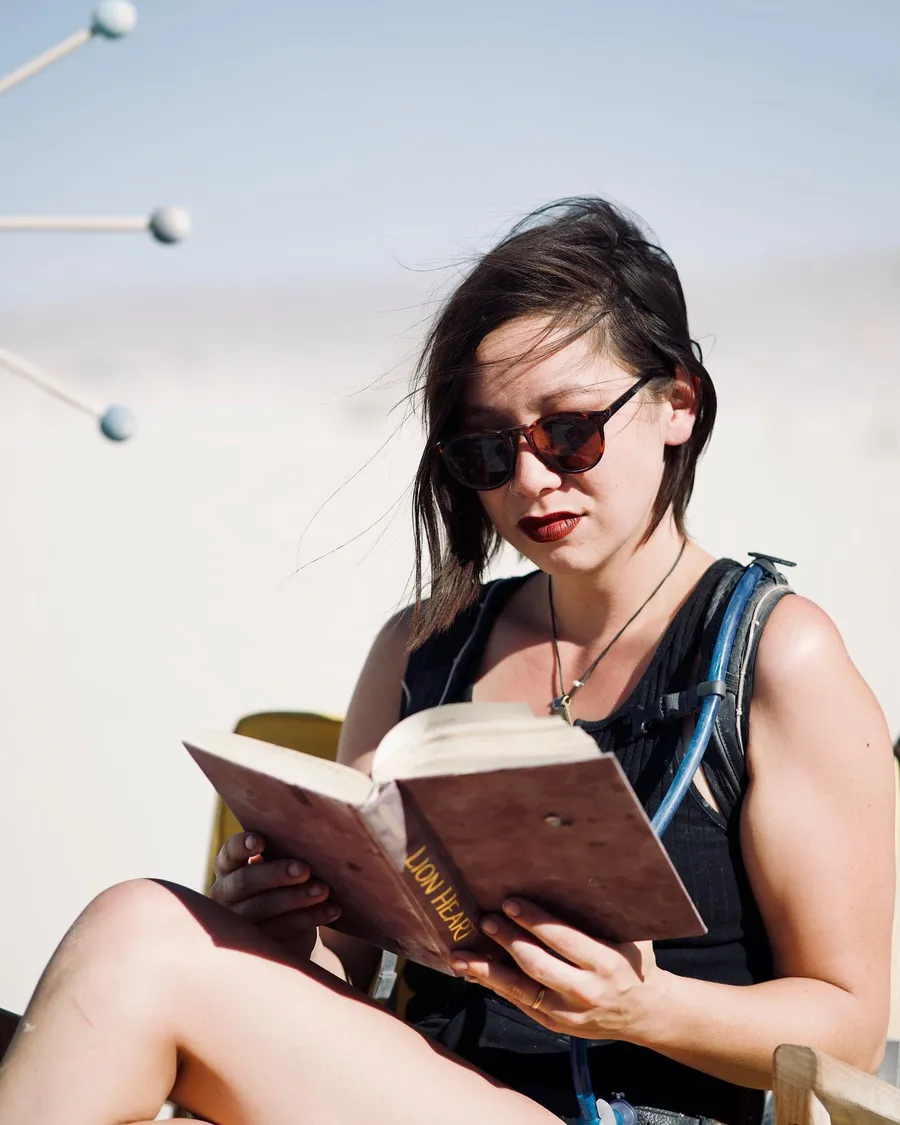 A seated person in sunglasses reads a hardcover book on the playa