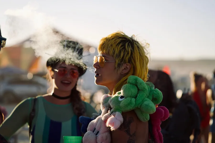 Two people in colorful costumes talk, one with short yellow hair and plush toys around their shoulders