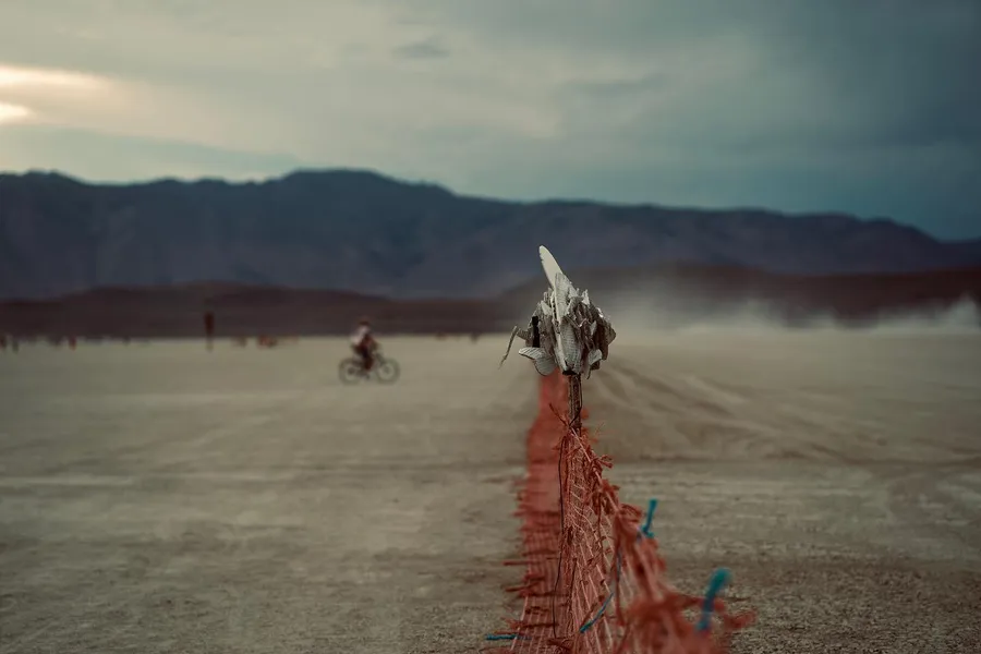 A ribbon fence leads toward a small horned sculpture, with a cyclist crossing the dusty playa beyond