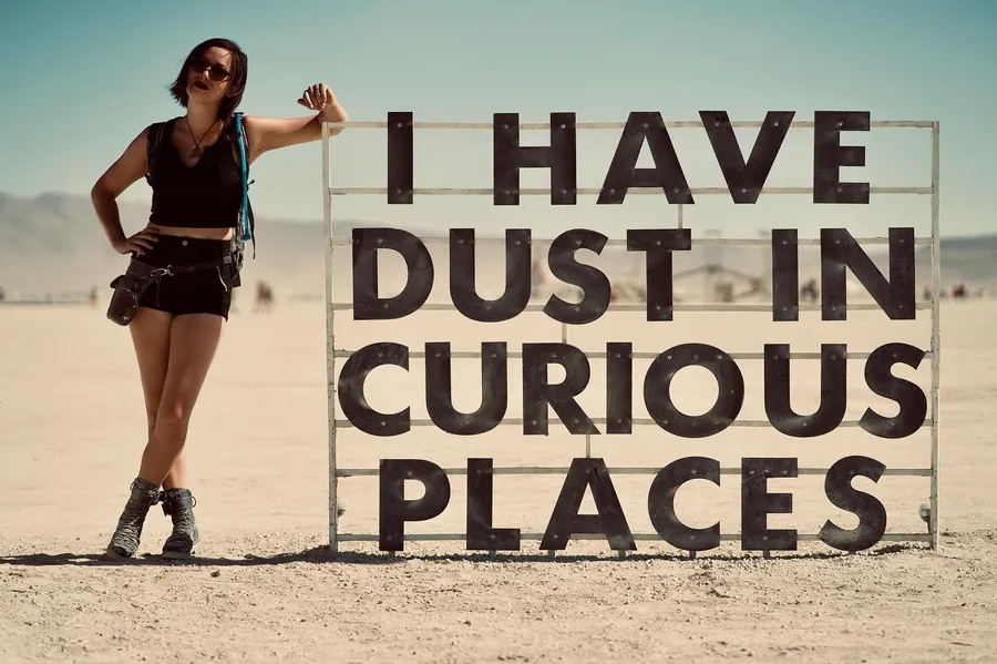 A person leans against a sign reading “I HAVE DUST IN CURIOUS PLACES”
