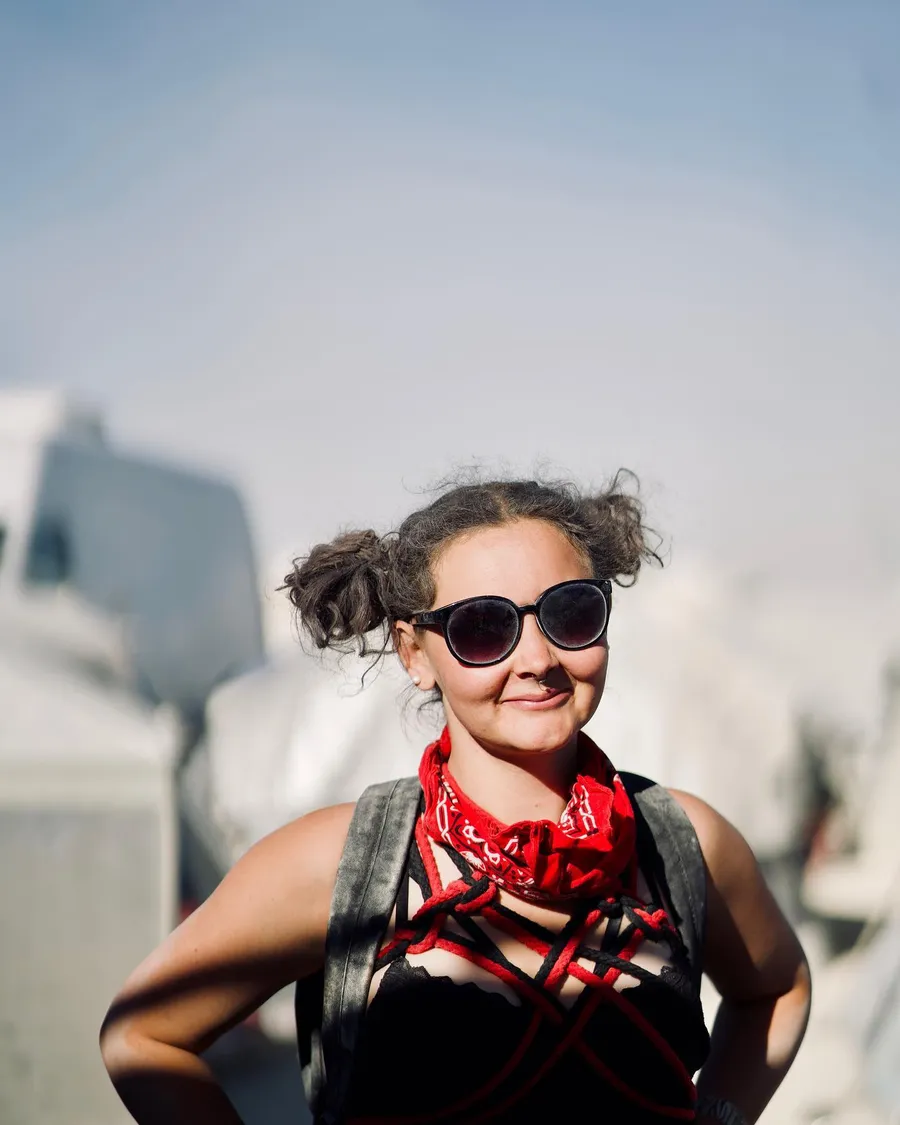 A smiling person with pigtails, round sunglasses, and a red scarf poses with hands on hips