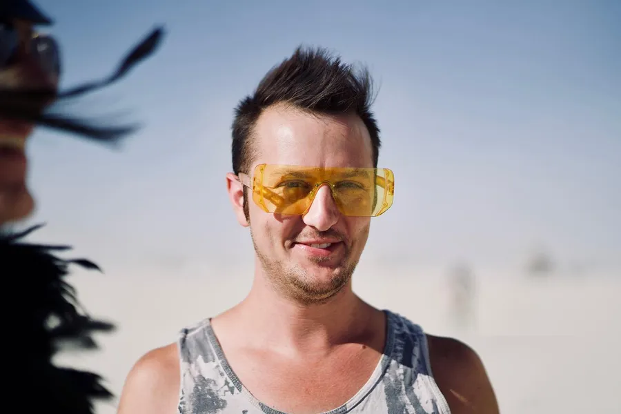 A smiling person wears oversized translucent yellow glasses on the playa