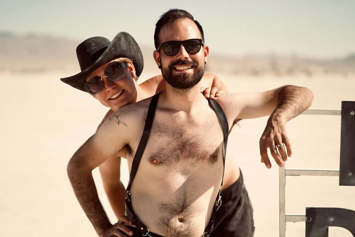 Two people in sunglasses pose together, one in a cowboy hat and one wearing suspenders