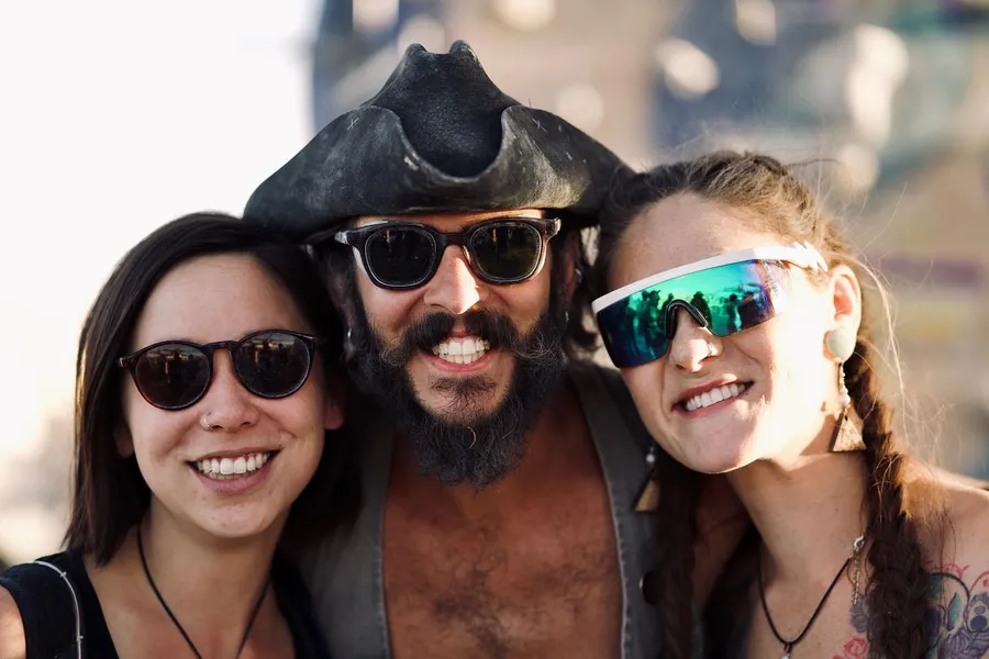 Three smiling people in sunglasses lean together, the center person wearing a pirate-style hat