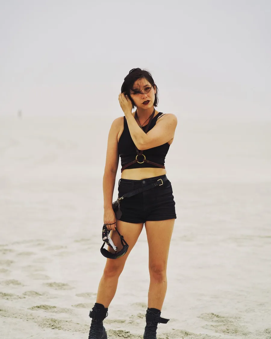 A person in black stands alone on the playa holding a camera and adjusting their hair