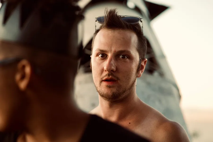 Close-up of a shirtless person with sunglasses on their head as another face enters the foreground