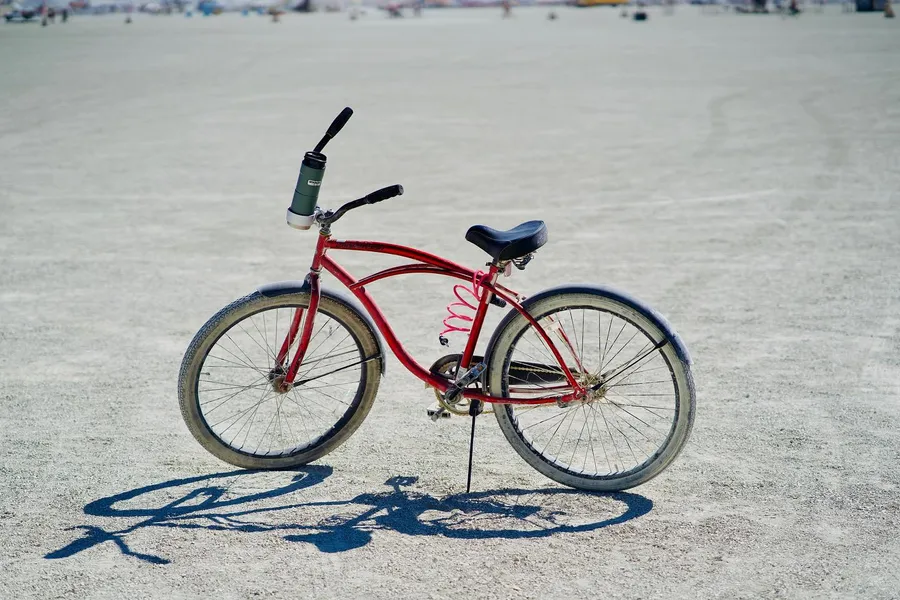 A red cruiser bicycle stands alone on the playa with a bottle mounted on its handlebar