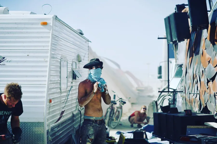A shirtless person in a cowboy hat holds a turquoise scarf over their face beside a trailer