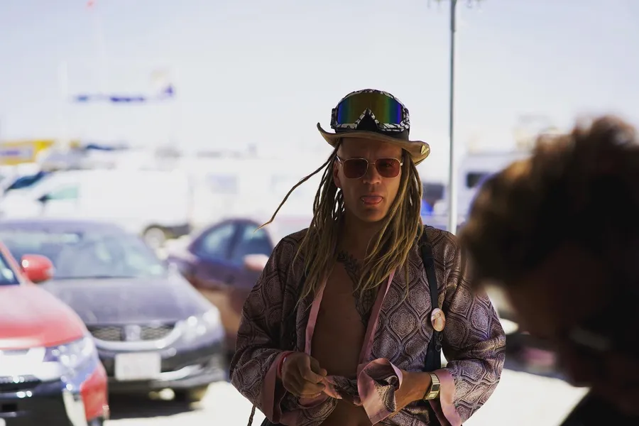 A long-haired person in sunglasses and a goggled top hat wears an open metallic-patterned coat