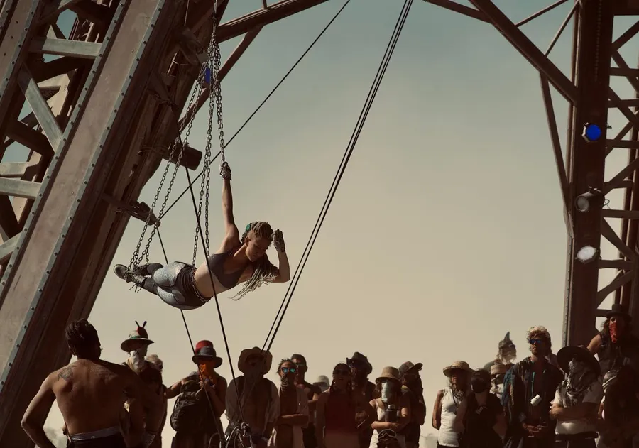A performer hangs sideways from chains beneath a large arched structure as a crowd watches