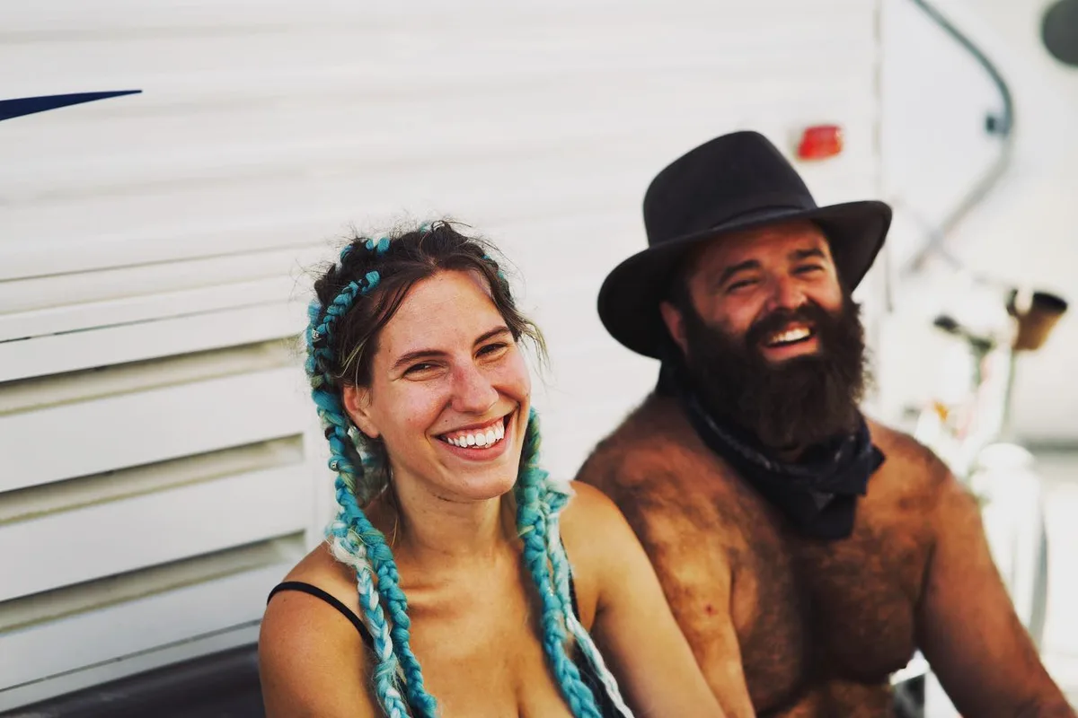 Two smiling people, one with turquoise braids and one bearded in a black hat, pose beside a trailer