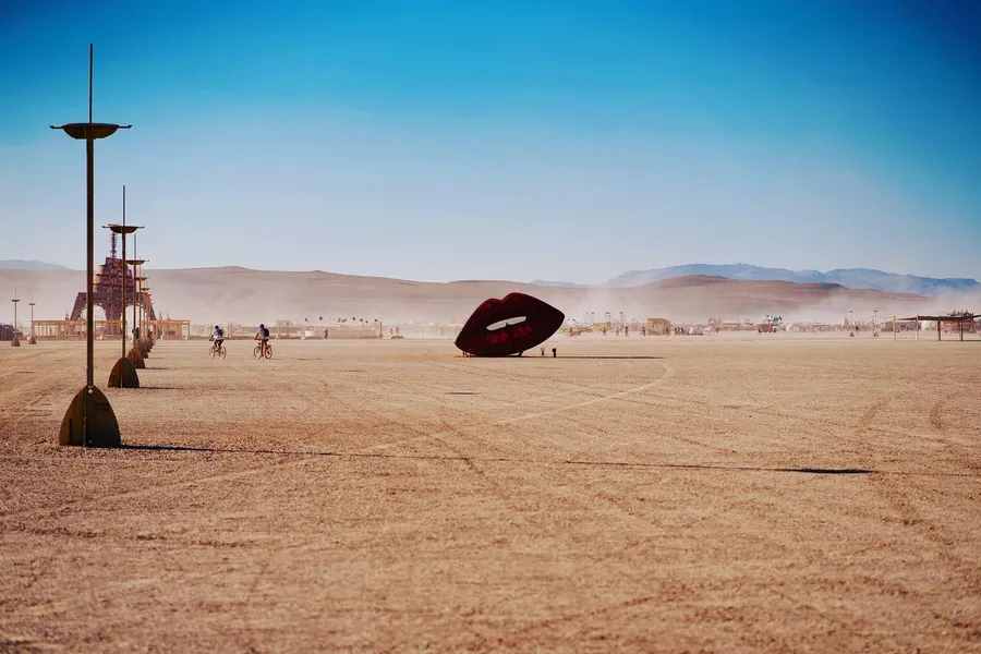 A giant black-and-red lips sculpture stands on the open playa beyond two cyclists