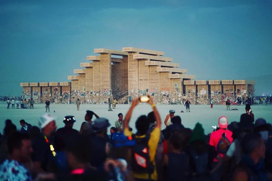 A crowd gathers before the stepped wooden Temple on the playa in daylight