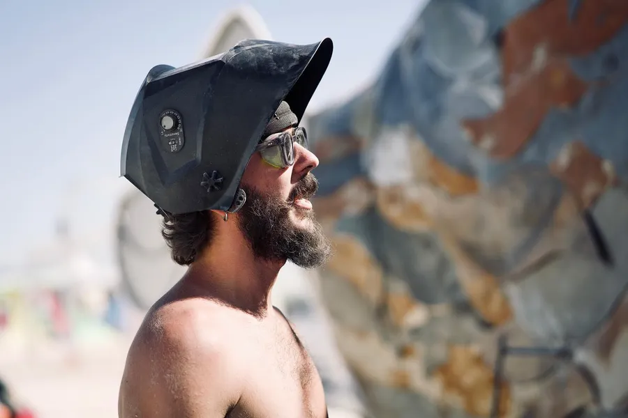 A shirtless bearded man in sunglasses looks up beneath a raised welding helmet