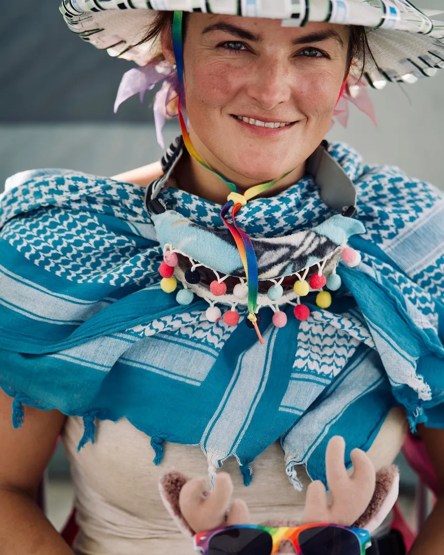 A smiling woman wears a patterned sun hat and a turquoise tasseled scarf
