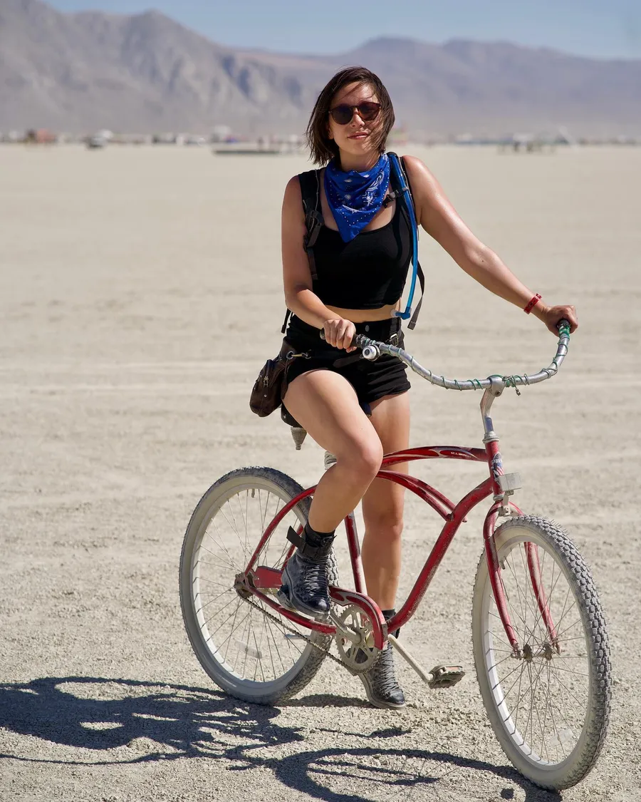 A woman in sunglasses and a blue bandana rides a red bicycle across the playa