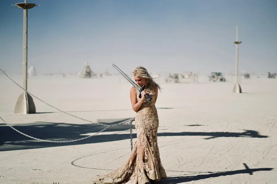A woman in a long gold lace gown stands beside chain barriers on the playa