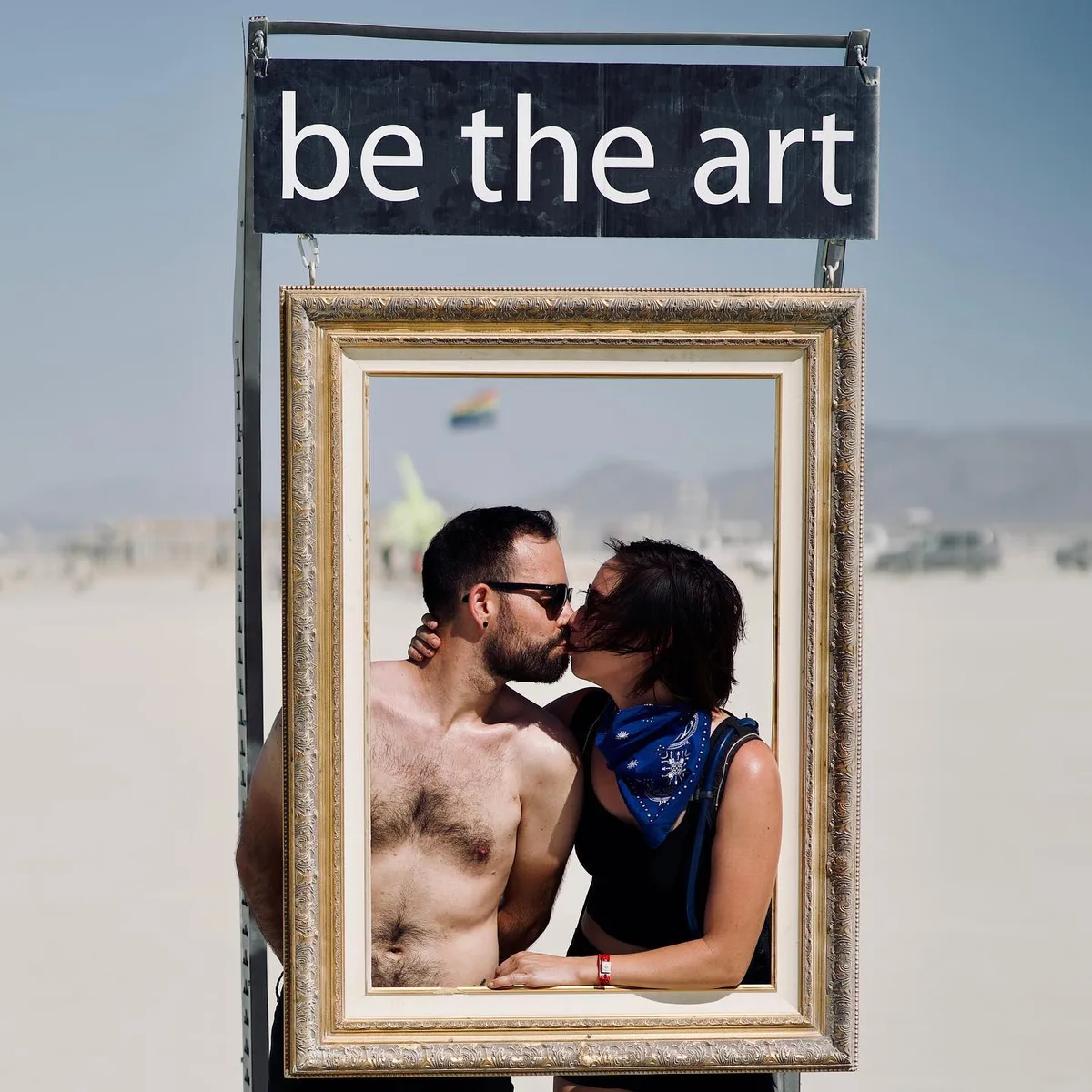 Two people kiss inside a gold picture frame beneath the words “be the art”