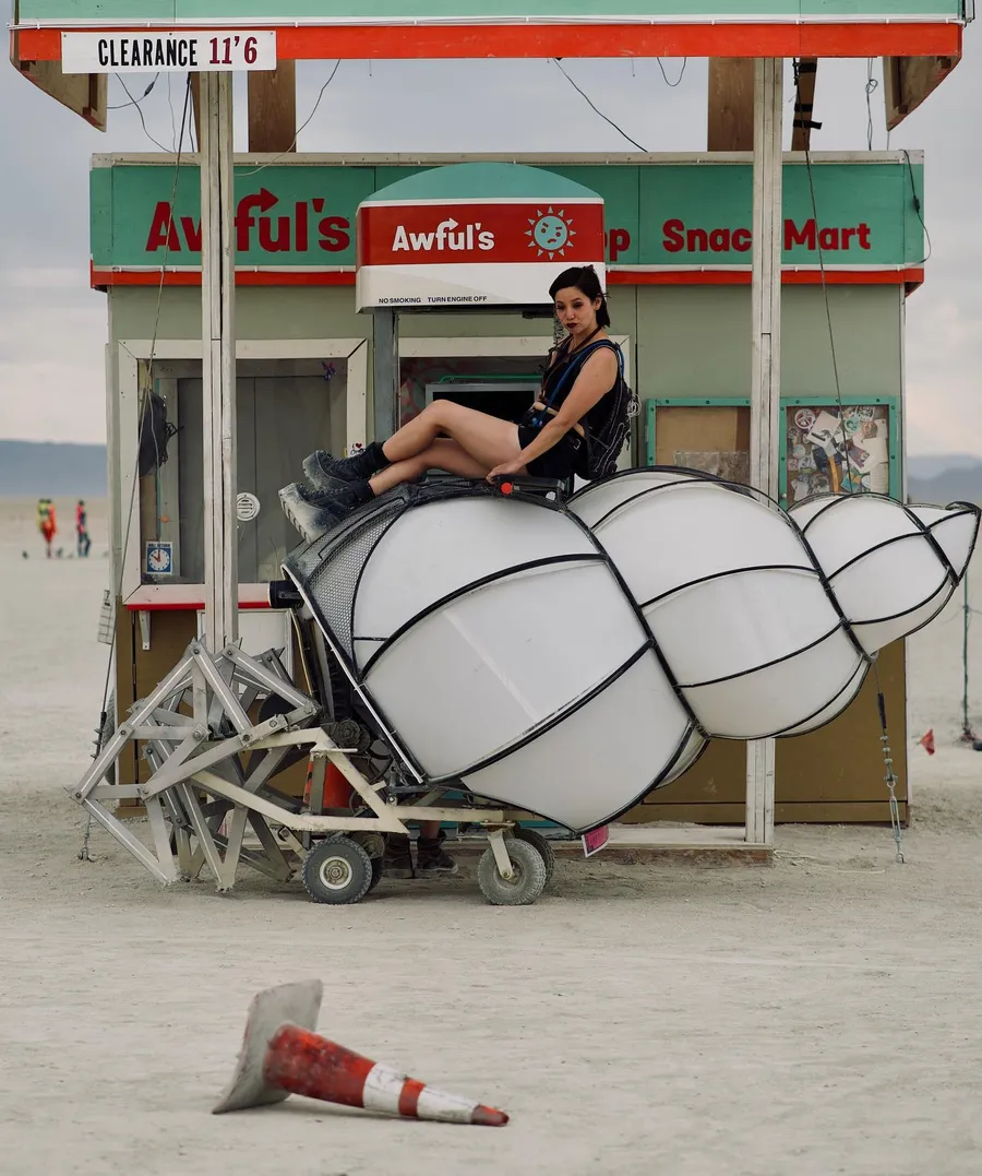 A woman sits atop a white segmented art vehicle in front of the Awful’s Stop Snack Mart installation