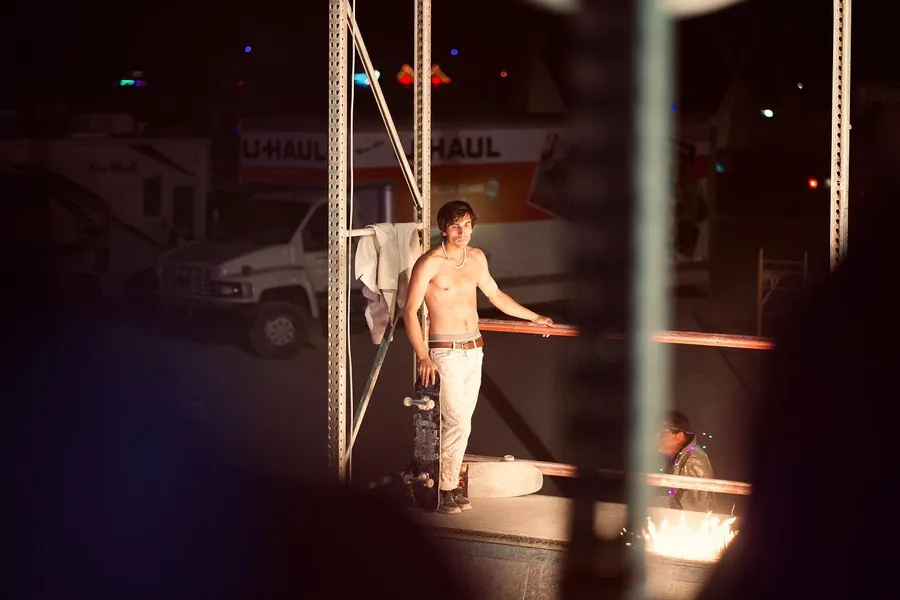 A shirtless skateboarder stands on a raised platform between fire bowls at night