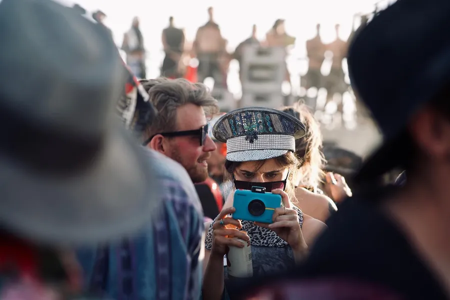 A person in a jeweled cap aims a turquoise instant camera through a crowd