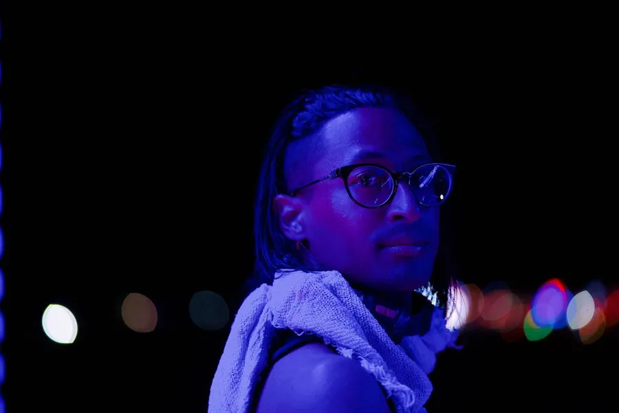 Night portrait of a person with glasses and short dreadlocks lit in blue and purple