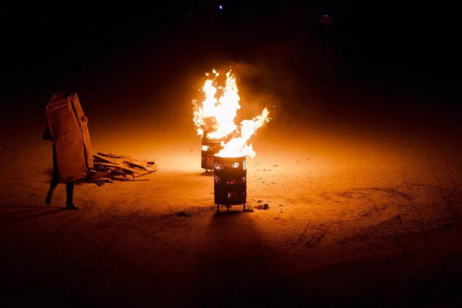 A person in a cardboard book costume watches two flaming barrels at night