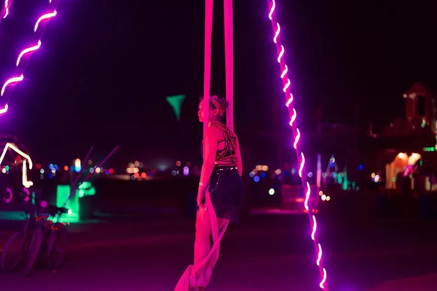 An aerialist holds pink silks between glowing ropes at night