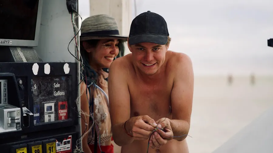 A shirtless man works with small wires beside an old gas pump while a woman smiles behind him