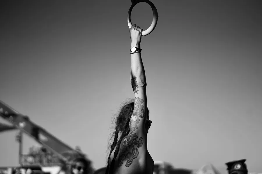 A tattooed woman reaches up to grasp a hanging ring in a black-and-white image