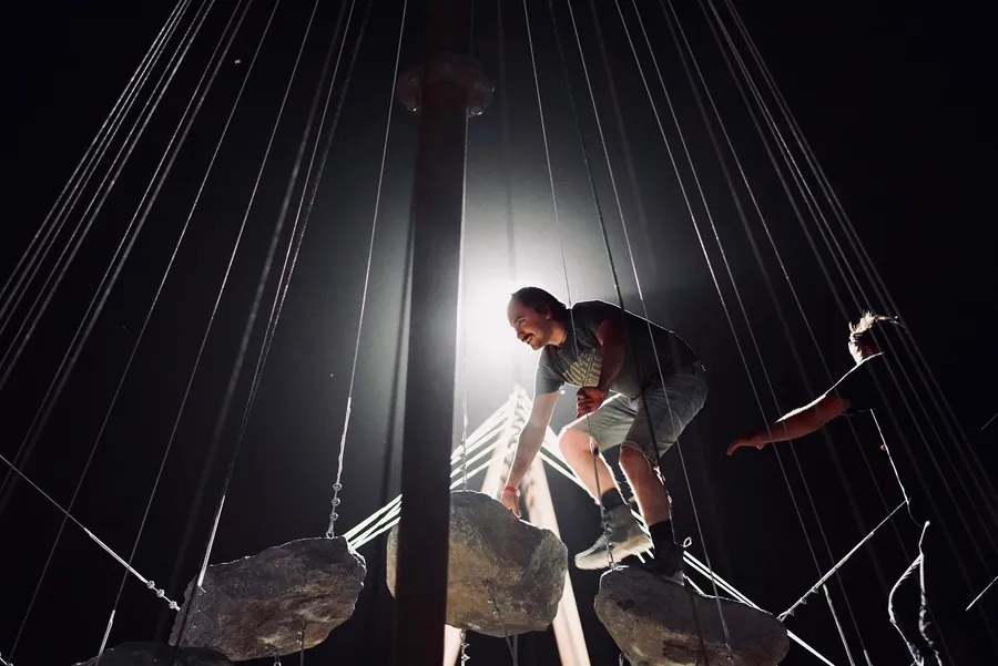 Two people climb among suspended rocks and cables, dramatically backlit at night