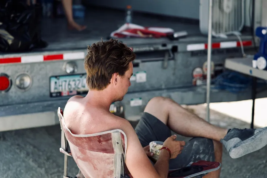 A shirtless person sits in a worn chair beside a trailer