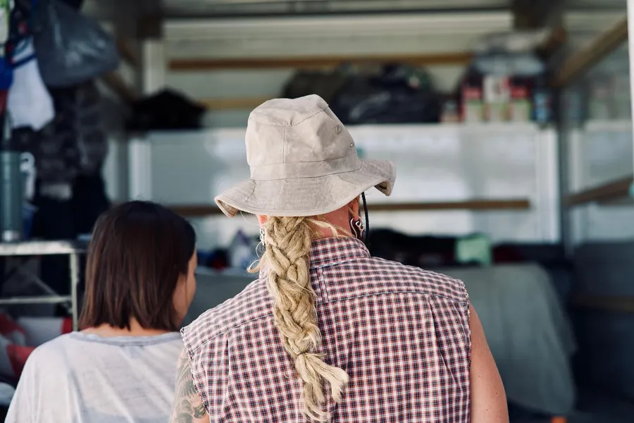 Two people seen from behind at a camp kitchen, one wearing a white hat with a long braid