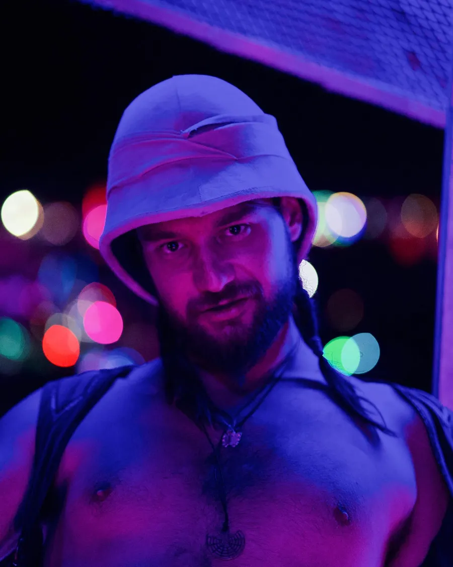 A shirtless man in a white hat is lit purple against colorful lights at night