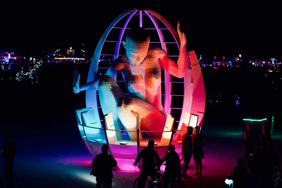 A giant seated human figure glows red and blue inside an egg-shaped frame as people gather below