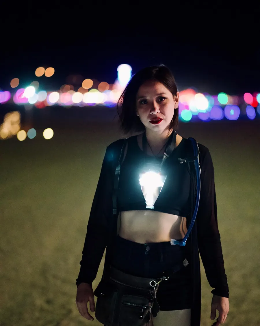 A woman in black wears a bright lightbulb-shaped lamp on her chest with playa lights blurred behind her