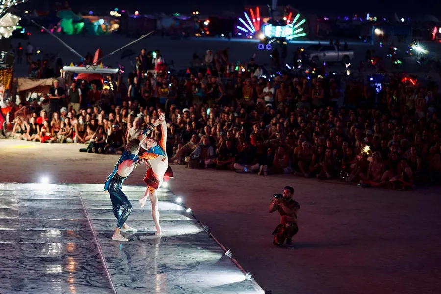 Two costumed dancers perform in a spotlight before a large nighttime crowd