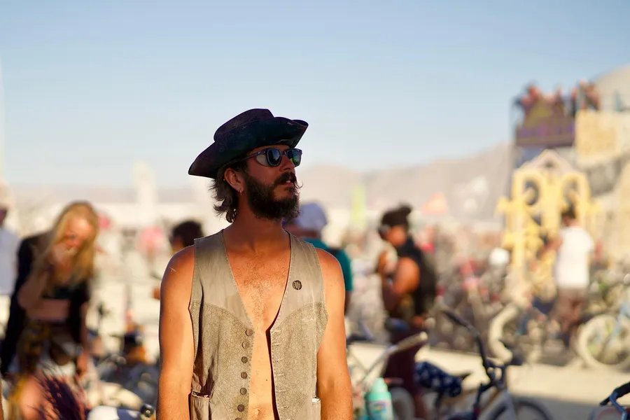 A bearded man in round sunglasses, a brimmed hat, and a vest stands amid bicycles and a crowd