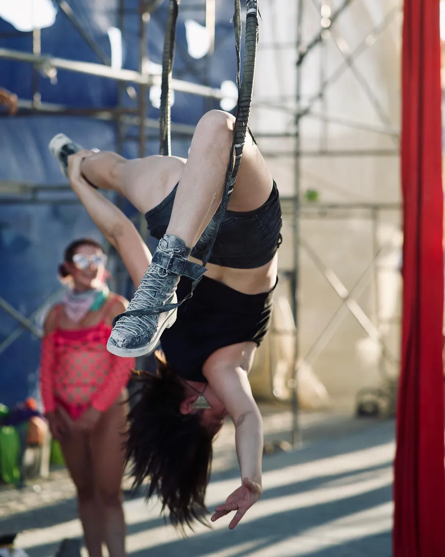 An aerialist hangs upside down from a hoop in a sunlit camp