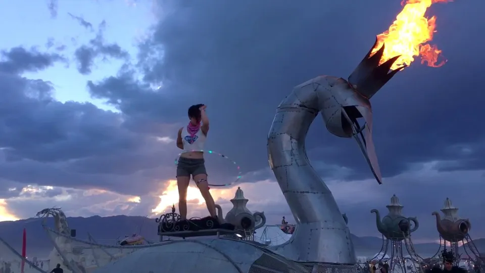A person stands atop The Prodigal Swan as its crown shoots flame at dusk