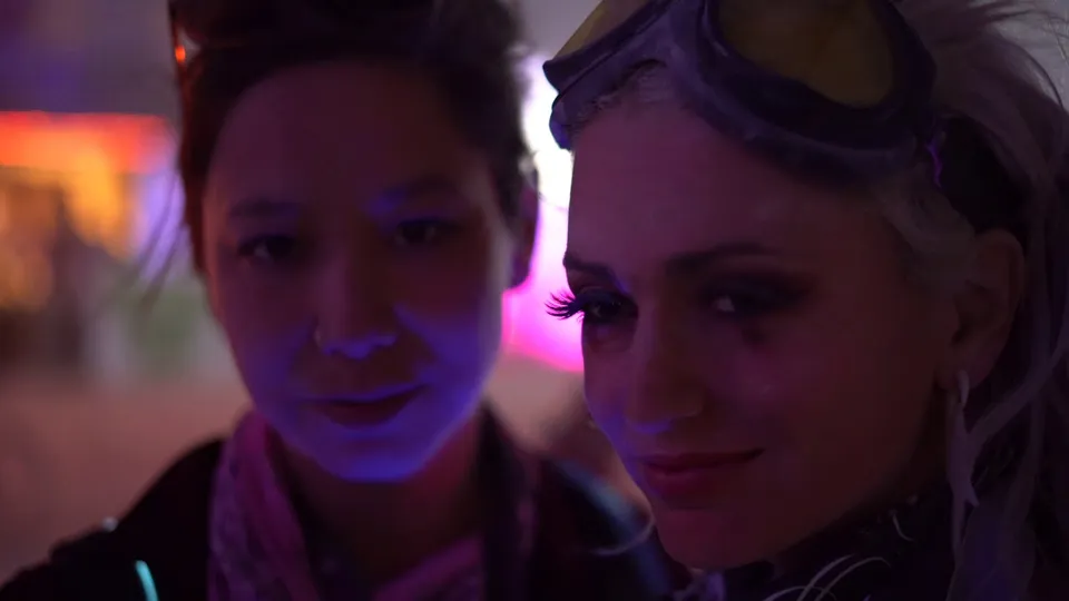 Two people pose close together under purple light, one wearing goggles