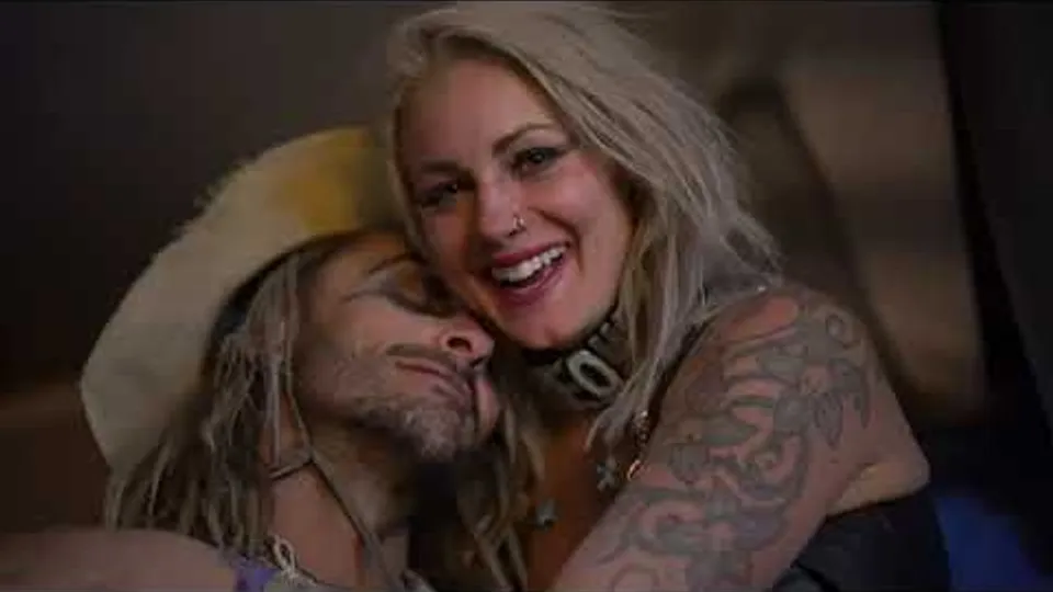 Two people embrace, one smiling toward the camera and the other wearing a brimmed hat