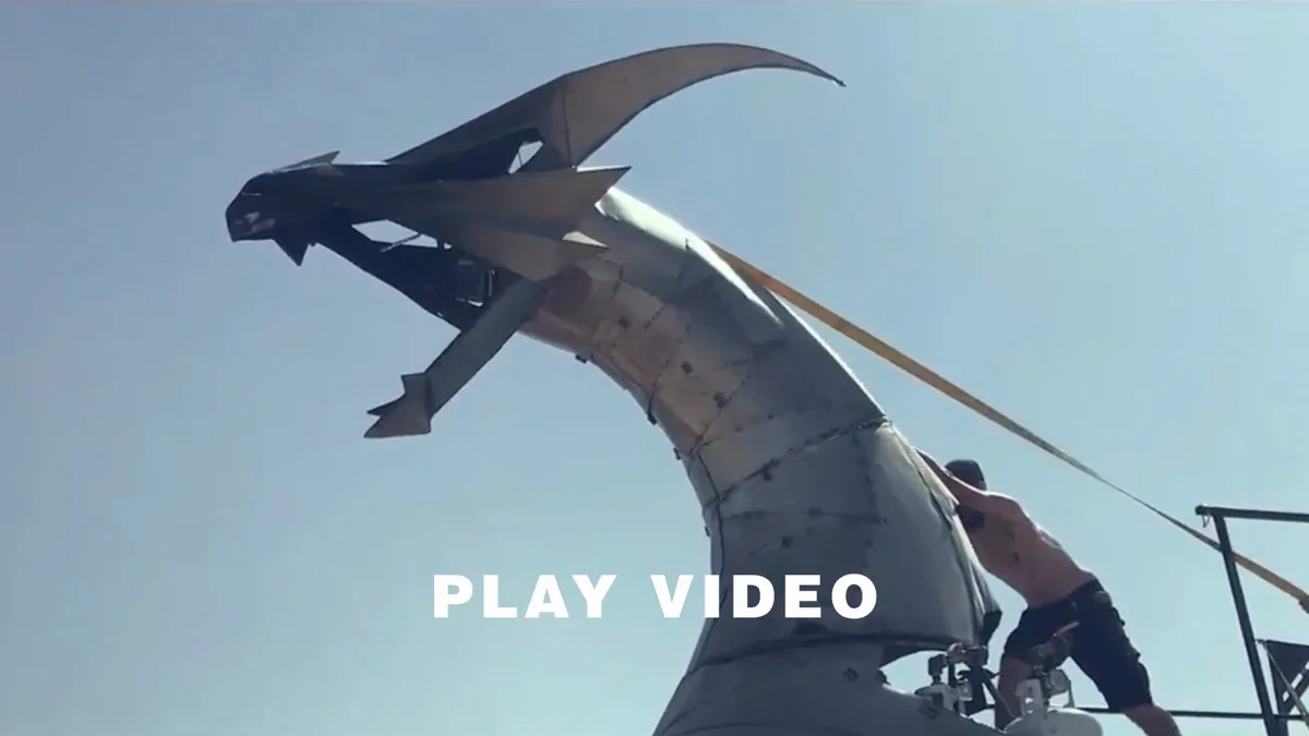 A person climbs beside Heavy Meta's steel dragon head beneath the words Play Video